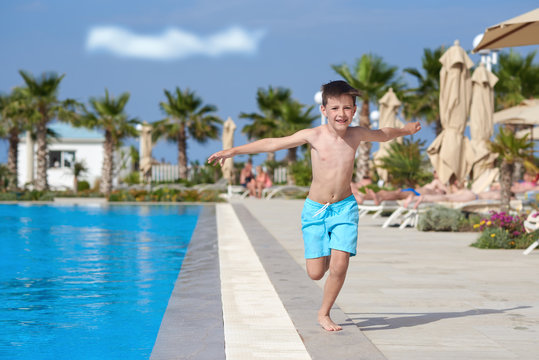 Portrait of Caucasian boy running along swimming pool at resort. He is smiling into camera and holds arms wide open.