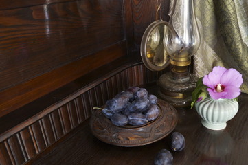 Rustic background with plums plate, gas lamp and hibiscus flower

