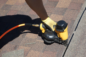 Roofer builder worker with nailgun installing Asphalt Shingles or Bitumen Tiles on a roof of a new...