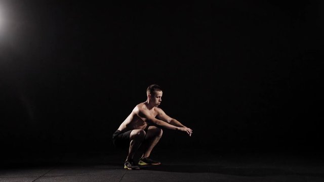 Fitness model doing jumping squats shirtless in a dark studio.