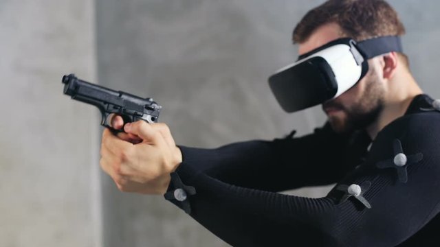 side view serious bearded man in motion-capture suit with sensors using vr headset hands holding weapon gun shooting then stop and looking around computer video-game entertainment industry animation