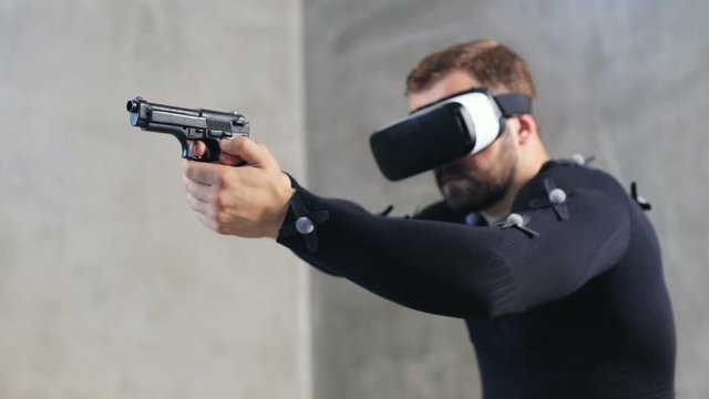 adult brunette man in motion capture suit with sensors wears vr glasses hands holding weapon gun shooting shoot video game entertainment industry console developers futuristic technology hi-tech