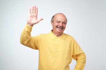 Naklejka premium Friendly-looking attractive european pensioner in yellow t-shirt waives hand in hello gesture while smiling cheerfully.