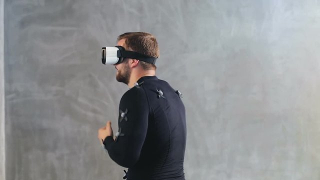 portrait of cheerful young man have fun dance while getting experience using 3D VR headset glasses of virtual reality in grey studio motion capture system suit with sensors developers testing program