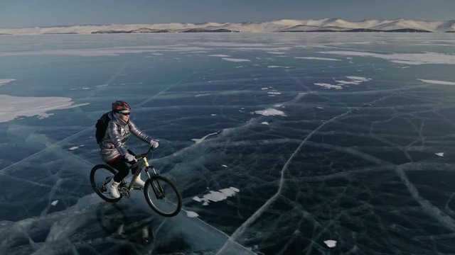 Woman is riding bicycle on the ice. The girl is dressed in a silvery down jacket, cycling backpack and helmet. Shooting with a quadrocopter drone. Ice of the frozen Lake Baikal. The tires on the