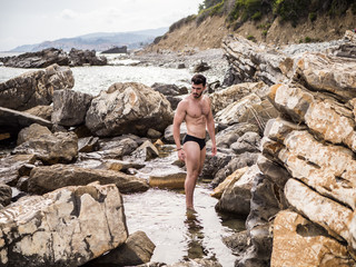 Handsome muscular young man standing on a rocky beach, relaxed, shirtless, looking away