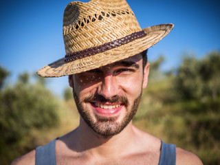 Attractive, fit young man relaxing walking in a grass field, wearing straw hat, looking at camera, smiling