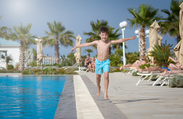 Smiling European boy running along swimming pool at resort in summer. He is holding arms wide open.