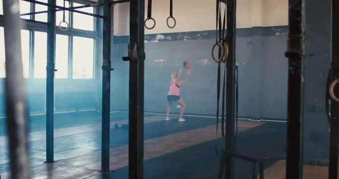 WIDE LR DOLLY Young adult sporty Caucasian woman doing exercise with heavy medicine ball in functional gym, morning shot. 4K UHD 60 FPS SLOW MOTION