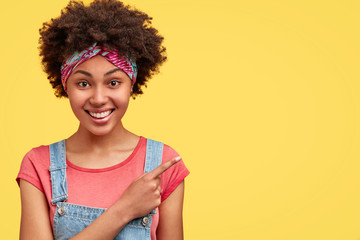 Curly dark skinned female with joyful expression, dressed casually, points at upper right corner, isolated over yellow background, suggests to visit this cafe. Positive African American woman indoor
