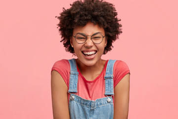 Happy African American female giggles and closes eyes, shruggs shoulders, dressed in casual t shirt...