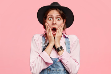 Photo of surprised female gardener opens mouth widely, keeps both hands on cheeks, reacts on something terrible, dressed in shirt, overalls, black hat, isolated over pink background. Omg, I am shocked