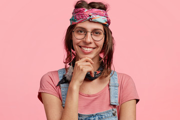 Cheerful young female hippy, holds chin, has toothy smile, wears fashionable headband and casual clothes, belongs to special subculture, isolated over pink background. People and emotions concept