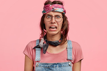 Displeased young female hippie with discontent expression, curves lips, sees something negative, dressed in casual t shirt, overalls and headband, isolated over pink studio wall. Negative emotions