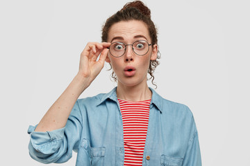 Surprised young female with freckled face, stares with shock, wears spectacles, being amazed, dressed in striped red sweater and denim shirt, witnessed shocking accident. Astonishment concept