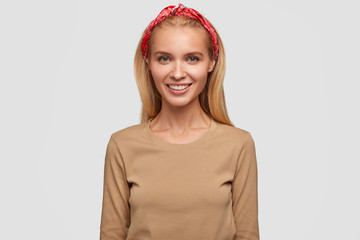 Young lovely blonde female has shinig smile, long hair, wears headband and beige casual sweater, expresses positive emotions, poses in studio, ready for date with boyfriend or meeting with friend