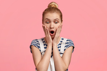 Emotive blonde female looks down with surprised expression, touches cheeks, notices something terrible and unexpected, wears striped casual t shirt and overalls, isolated over pink background