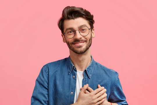 Pleased kind hearted bearded young male keeps both palms on chest, looks positively at camera, expresses truthful feelings to girlfriend, wears stylish denim shirt, isolated over pink background.