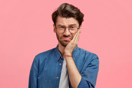 Image of displeased bearded young guy has sullen facial expression, touches cheek with hand, dressed in fashionable denim shirt, poses against pink background, being dissatisfied with something