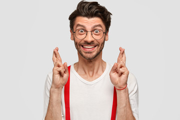 People, body language and expectation concept. Glad Caucasian young male with stubble, keeps fingers crossed, wears white shirt with red braces, has happy expression, isolated over studio wall