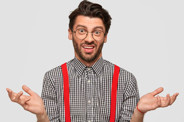 Clueless hesitant male with trendy haircut, wears fashionable outfit and spectacles, shruggs...