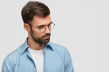 Photo of unshaven young male with dark stubble and hair, wears spectacles and shirt, focused aside, poses against white background with copy space for your advertisement. Thoughtful hipster.