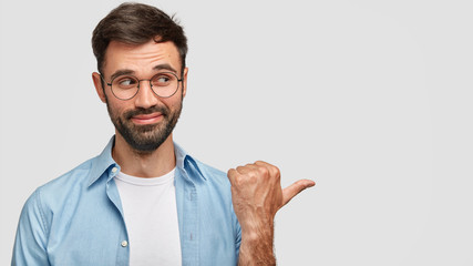 Portrait of attractive unshaven male with dark stubble, points aside with thumb, shows blank space...