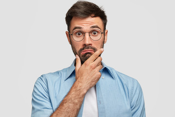 Photo of surprised bearded male holds chin and looks in hesitation, wonders latest news, wears...