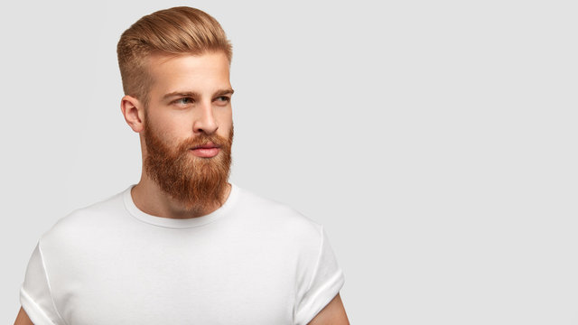 Serious thoughtful male with ginger beard, dressed casually, focused somewhere, isolated over white background with free space on right for your advertising content. Pensive red haired hipster