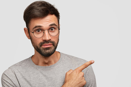 Photo of handsome European male with thick beard and mustache, points with fore finger aside at free space for your promotional content, wears round spectacles and grey t shirt. Advertisement