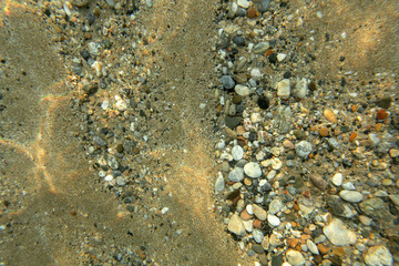 Underwater photo - sand sea bottom with small pebbles and stones, lit by sun, shot from above. Abstract marine background.