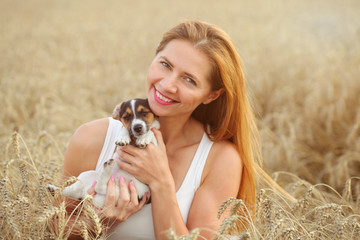 Young woman in white dress, sitting in the wheat field, holding Jack Russell terrier puppy