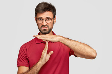 Photo of displeased bearded male keeps hands perpendicularly, has uncertain expression, tries to explain route or direction, wears round spectacles and red t shirt, isolated over white background