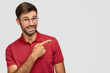 Indoor shot of positive bearded male in casual red t shirt, points with index finger aside, shows...