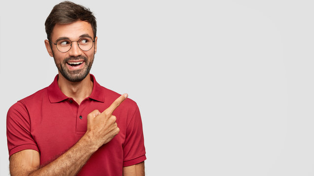 Indoor shot of glad unshaven man with thick beard and mustche, points with index finger at free space for your advertisement, dressed casually, being in high spirit, smiles broadly, has white teeth
