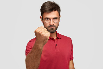Serious angry male shows fist, ready for fight or challenge, has stern expression, wears casual red...