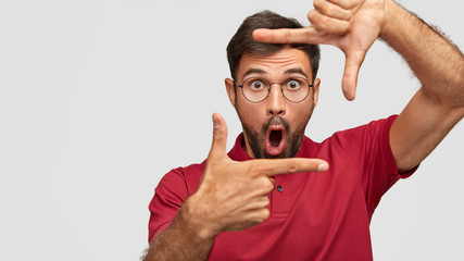 Horizontal shot of bearded stunned male stares through frame formed by hands, has unexpected...