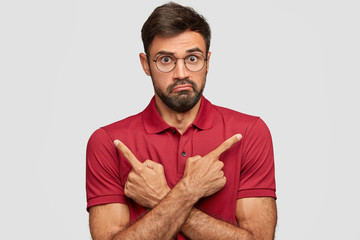 Studio shot of confused clueless male with beard and mustache, points with index fingers at different directions, has unaware expression, surprised look, poses against white background, gestures