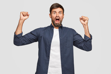 Puzzled crazy male with frightened expression, yells loudly and desperately, keeps hands clenched in fists, has terrific look, dressed casually, isolated over white wall. People and reaction concept