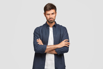 Waist up shot of serious unshaven male with confident expression, keeps arms folded, listens necessary information attentively, wears stylish shirt, poses against white studio wall. People, lifestyle