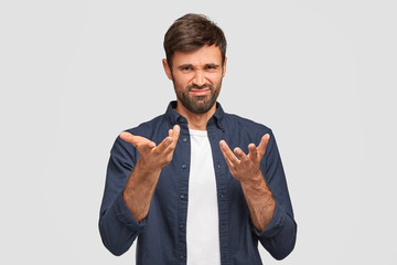 Indoor shot of bearded male frowns face, has displeased expression, gestures with hands, dressed in fashionable clothes, isolated over white background. Attractive young European man has upset look
