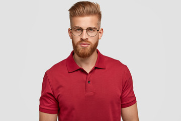 Candid shot of self confident serious Caucasian male with foxy hair and beard, dressed casually, looks directly at camera with mysterious expression, wears spectacles, isolated over white background