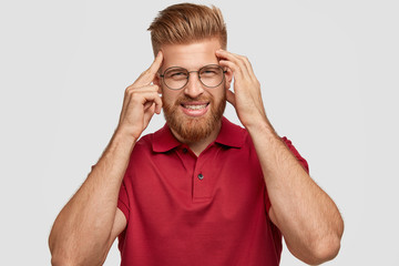 Fototapeta premium Photo of cheerful red haired young hipster has bad memory, keeps hands on temples, tries to remember something, has friendly smile, wears casual outfit, stands alone against white background.