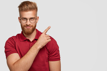 Studio shot of attractive young ginger male with thick beard, points at upper right corner, has...