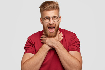 Photo of tired bearded male with negative facial expression, suffers from suffocation, keeps hands on neck, looks in displeasure at camera, has ginger hair, dressed casually, isolated on white wall