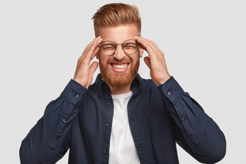 Horizontal shot of attractive stressful foxy male clenches teeth, keeps hands on temples, has headache, dressed in fashionable clothes, isolated over white background. People, emotions and feeling