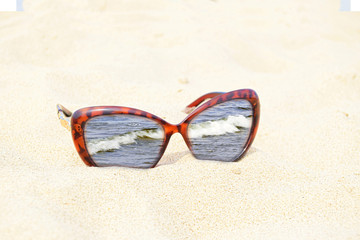 Sunglasses are on sand. The image of the sea is in a frame of sunglasses. Glass lenses are inserted into a casing of color of a tiger.