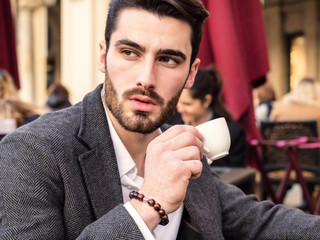 Handsome young man drinking espresso coffee, wearing elegant coat posing at table outside on elegant urban background in Turin, Italy