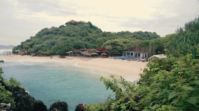 White sandy beach with straw umbrellas, houses and boats near green hill in Java, Indonesia