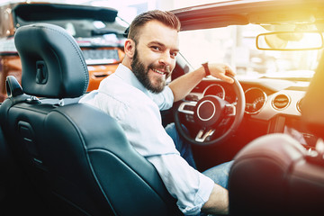 Handsome bearded man choosing a new sport cabriolet car in the dealership. Guy looking on camera